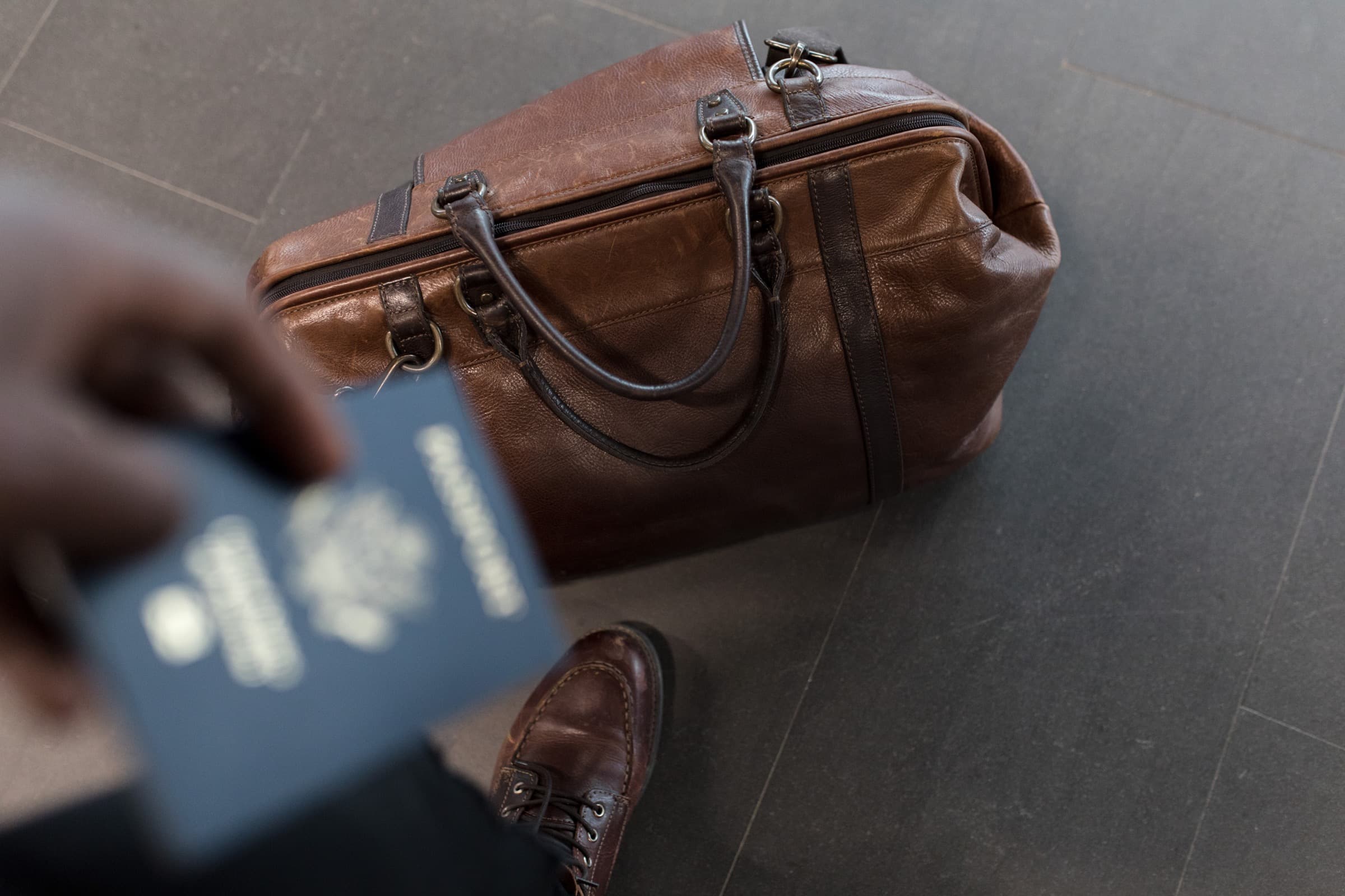 A traveler holding a passport beside a brown leather travel bag