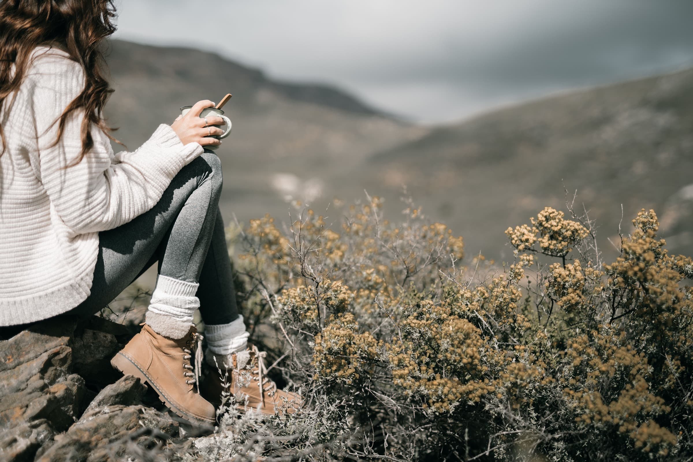A person in hiking boots holding a cup while overlooking a mountain landscape